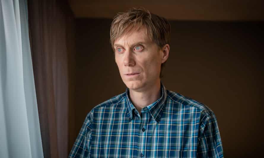 A pale man with floppy hair wearing a plaid shirt stares unsettlingly out of a window