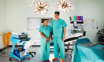 Lucy Khan and Chris Cartlidge, standing side by side wearing scrubs, in an operating theatre.
