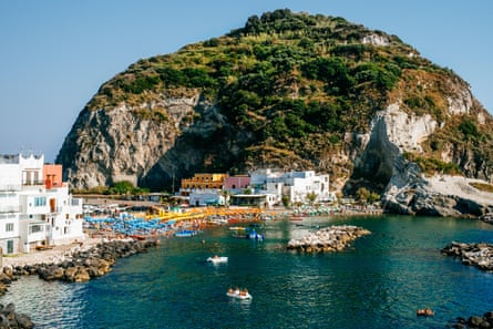 A crowded beach on the island of Ischia, Italy, between a large rock formation and a small town.