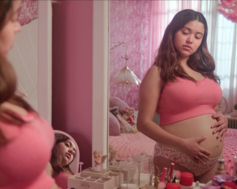 A pregnant woman in a pink crop top looks at her reflection in a mirror in a pink-decorated bedroom