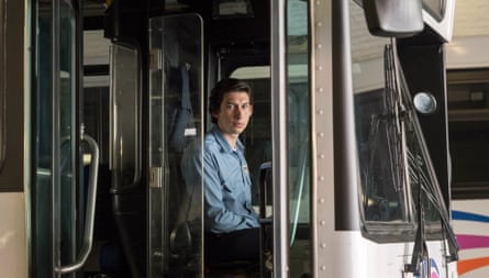 Playing the role of a bus driver, he sits in the front seat of a bus, looking at the camera to his right