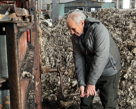 A man holds a crank handle on a piece of machinery. He is in a large shed that appears to be full of wool