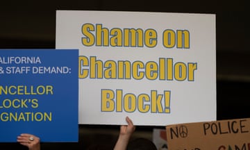 A hand holds up a white posterboard sign that says in yellow letters Shame on Chancellor Block, amid other signs.