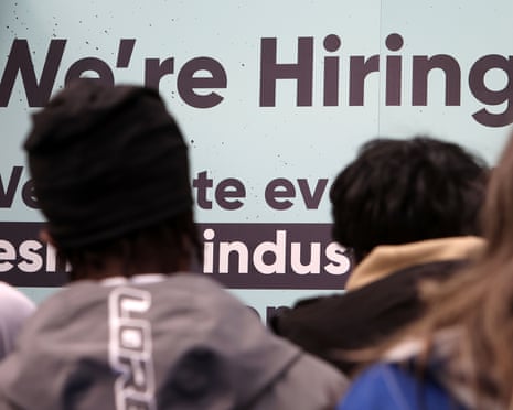 People stand facing a large sign that reads "We're Hiring" at a job fair