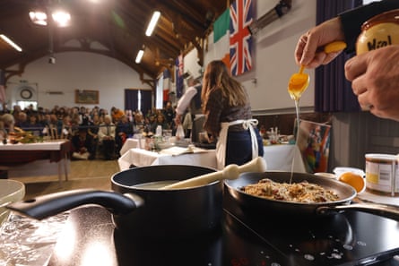 A competitor spoons honey into a pan of oats on a hob. Spectators sit in rows at the back of the hall.