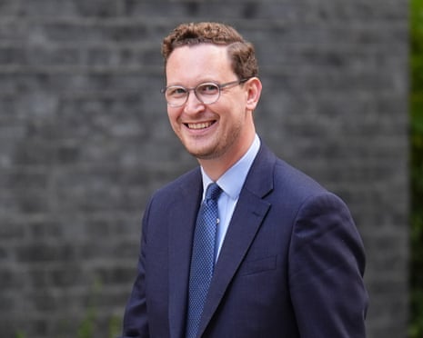 Darren Jones, in a navy suit, light blue shirt and glasses, stands outdoors against a blurred stone wall