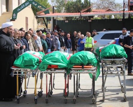 four bodies on trolleys wrapped in green fabric attended by religious figure and mourners.