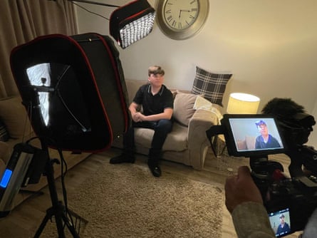 Charlie Simpson is filmed as he sits on a beige sofa in a living room; he is spotlit under a professional-looking lamp and a person in the foreground is looking at him through the viewfinder of a video camera. Charlie wears a black polo shirt and dark trousers.