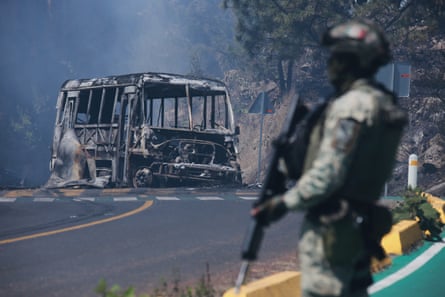 A soldier stands holding their rifle at the side of the pic, as a charred bus sits on the road in the background