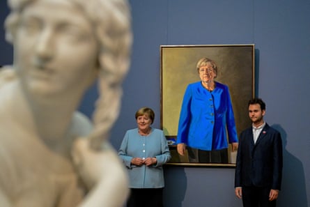 Merket and Queyras stand in front of the portrait, which is hung on a blueish-slate grey wall. There is a classical statue in the foreground of the picture.