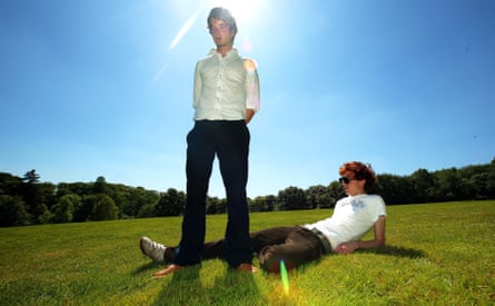 One man stands on grass while a man next to him lies down.