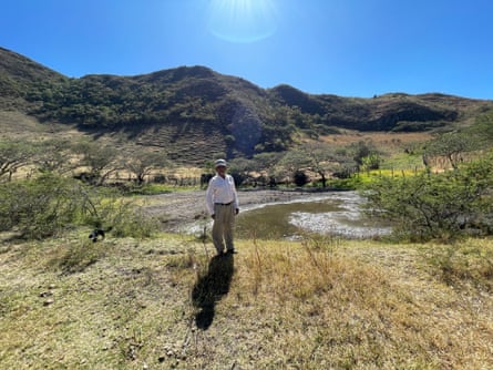 A man stands next to a small body of water among green hills