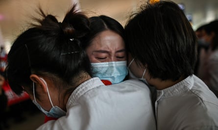 Three women in masks embrace, one facing towards the camera and two away