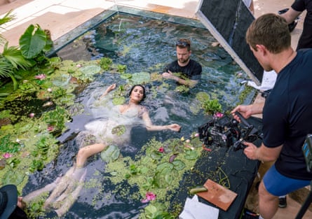 Padma Lakshmi lying in a pool with vegetation floating on top, being photographed.