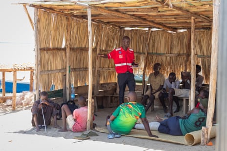 A group of men sit around a standing man doing a speech in a bamboo hut