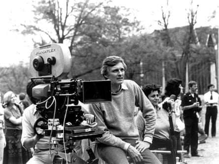 black and white photo of Alan Alda sitting in a sweater with a film camera to his right and people in the background