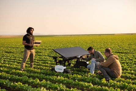 Tres personas están de pie o en cuclillas alrededor de un robot agrícola alimentado con energía solar en un gran campo de hierba.