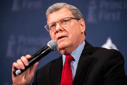 An older white man wearing glasses and a suit  speaks into a microphone he holds with one hand.