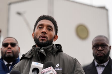 A man speaks into microphones outdoors, flanked by two other men.