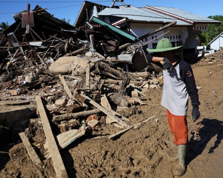 A man raises an arm to his face as he stands in mud beside a destroyed house