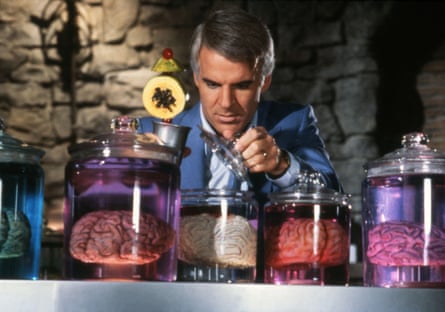 Steve Martin in a blue suit sitting in front of four jars with brains in them, and taking the lid off one of them