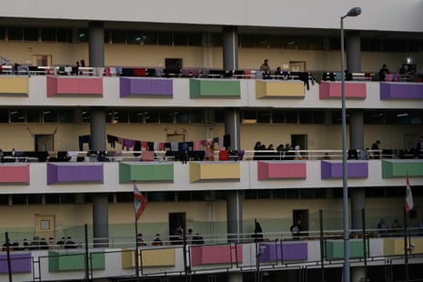 A wide shot of three long balconies of a school that are decorated with bright hyellow, pink, purple and green tiles. People are crowded the balconies and clothes lines heavy with drying clothes are visible.