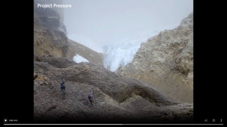 Two men standing at the moot of a section of brown mountain terrain with a small white glacier in the background