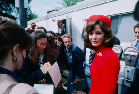 A woman in a red pillbox hat and red jacket surrounded by journalists