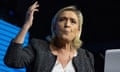 Marine Le Pen delivers a speech during a campaign meeting, 24 May 2024: she is seen in a head and shoulders photo with one hand raised as she talks. She is wearing a black collarless jacket over a white top with a gold necklace, and is seen against a black backdrop with an illuminated blue section.