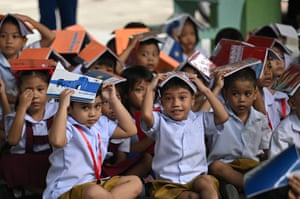 Young children wearing blue shirts and brown shorts sit on the floor covering their heads with their notebooks
