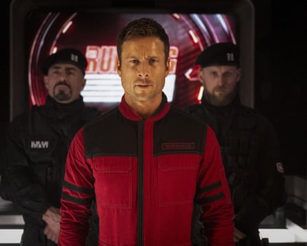 A shot of Glen Powell as Ben Richards in the 2025 film The Running Man. He stands front and centre, wearing a red and black tactical jumpsuit with a high collar. He has a serious, intense expression