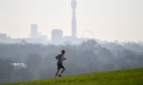 A man running on a green grass hill in the foreground, and blurry outline of trees and buildings in the background