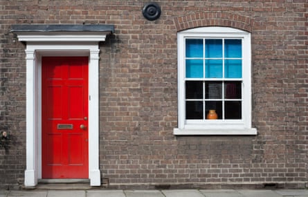 A red door, a window with blue blinds, and white surrounds on a brick town house in a British village.