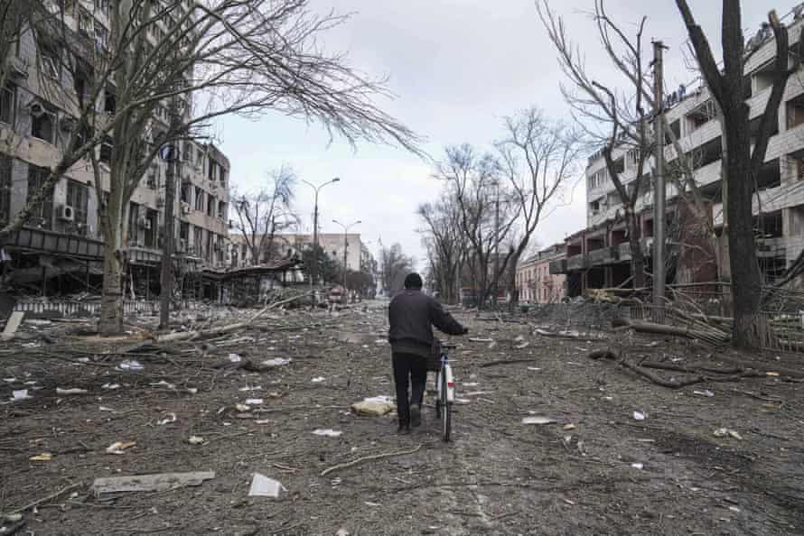 A man walks with a bicycle in a street with heavily bomb-damaged buildings and blasted trees.