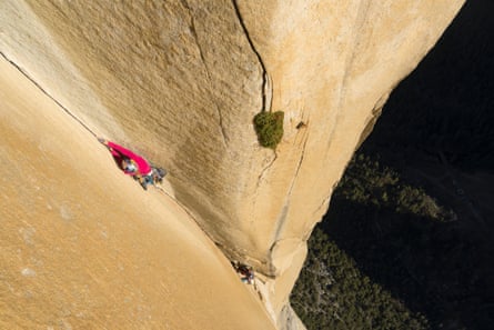 Woman in red climbing a sheer sandstone-colored rock face, with a male climber below her, and the ground thousands of feet below them both.