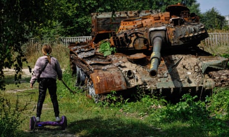 A teenager rides a hoverboard past a rusted tank in a field