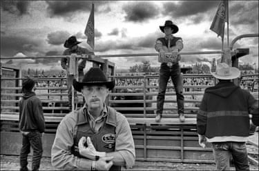 Cowboys wait near rodeo pens, one in a black hat holding gloves, before competing