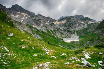 A grassy mountain valley with rocky peaks above on a cloudy day.