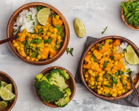 A photograph of a chickpea curry with rice, in a wooden bowl, with smaller bowls nearby containing broccoli and lime slices