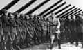 Black and white photo of Benito Mussolini in military gear marching past a row of soldiers