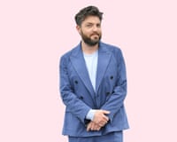 Tom Burke wearing a cornflour-blue unbuttoned suit over a pale-blue, long-sleeved top, standing with his hands clasped in front of him against a pale-pink background