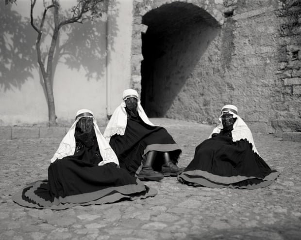 Three women wearing black cat masks with white scarves, sitting on the ground in a courtyard with their skirts arranged in a circle around them