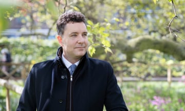 Edward Timpson stands in a park with new leaves and flowers, leaning on a wooden rail on the top of a wire fence.