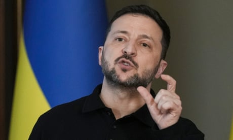 A head and shoulders photo of Volodymyr Zelenskyy as he speaks and gestures with a Ukrainian flag draped behind him