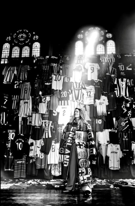 A woman stands in a room with the sun streaming in through high stained glass windows. She is surrounded by England football shirts and is holding a collection of England scarves