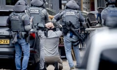 A man kneels on the ground with his hands behind his head and armed officers standing around him