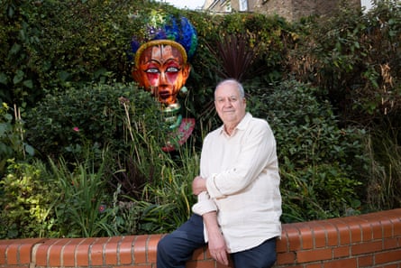 he wears a large white shirt, blue jeans and sits on a brick wall in a garden with lush foliage; a carnival mask is in the bushes behind him
