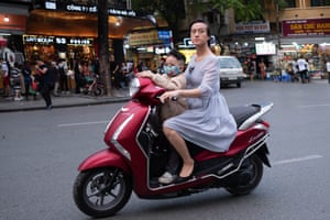 A woman wearing a dress driving a moped while a young boy sits in front wearing a facemask