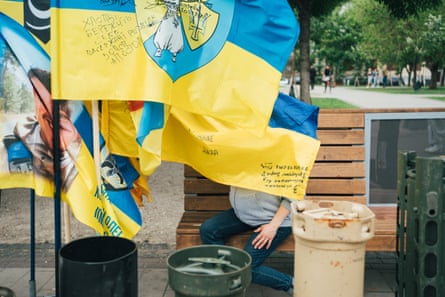 Yulia, obscured by fluttering Ukrainian flags, sitting on a bench in a Kyiv park.