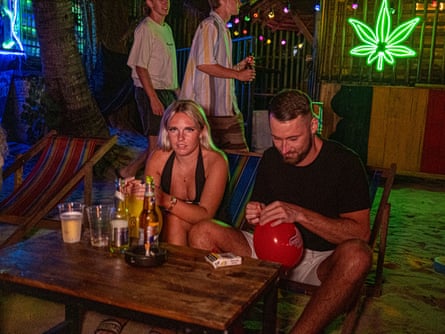 A man and woman sit at a low table holding balloons. A neon sign of a cannabis leaf is in the background